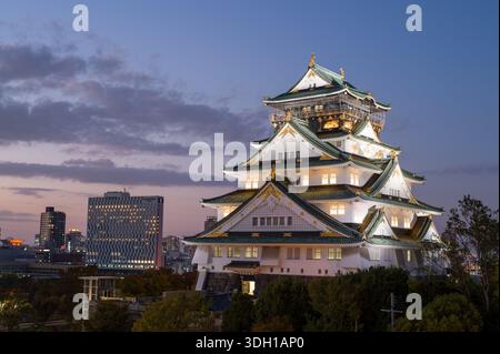 Osaka, Japan - 7. November 2023: Der Hauptturm der Burg Osaka wird in der Dämmerung beleuchtet, mit modernen Stadtgebäuden und Bäumen im Hintergrund unter teilweise bewölktem Himmel. Stockfoto