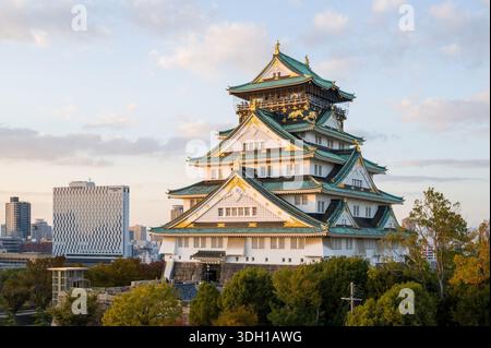 Osaka, Japan - 7. November 2023: Der Hauptturm der Burg Osaka mit seinem mehrstöckigen Dach mit grünen Fliesen und goldenen Akzenten ist bei Sonnenuntergang zu sehen, mit modernen Stadtgebäuden und Bäumen im Hintergrund. Stockfoto