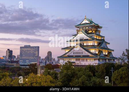 Osaka, Japan - 7. November 2023: Der Hauptturm der Burg Osaka wird in der Abenddämmerung beleuchtet, mit modernen Stadtgebäuden und Bäumen im Hintergrund unter teilweise bewölktem Himmel. Stockfoto