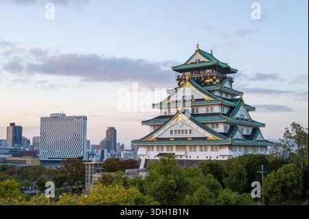 Osaka, Japan - 7. November 2023: Der Hauptturm der Burg Osaka mit seinem grün gekachelten Dach und goldenen Akzenten ist in der Abenddämmerung zu sehen, mit modernen Stadtgebäuden und Bäumen im Hintergrund. Stockfoto