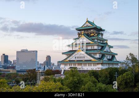 Osaka, Japan - 7. November 2023: Der Hauptturm der Burg Osaka mit seinem grün gekachelten mehrstöckigen Dach und Goldakzenten ist in der Abenddämmerung zu sehen, mit modernen Stadtgebäuden und Bäumen im Vorder- und Hintergrund unter teilweise bewölktem Himmel. Stockfoto