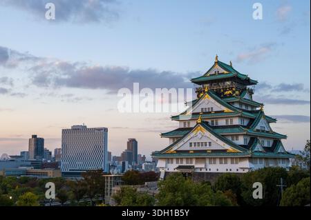Osaka, Japan - 7. November 2023: Der Hauptturm der Burg Osaka mit seinem grün gekachelten mehrstöckigen Dach und Goldakzenten ist in der Abenddämmerung zu sehen, mit modernen Stadtgebäuden und Bäumen im Hintergrund unter teilweise bewölktem Himmel. Stockfoto