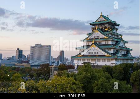 Osaka, Japan - 7. November 2023: Der Hauptturm der Burg Osaka mit seinem grün gekachelten mehrstöckigen Dach und Goldakzenten ist in der Abenddämmerung zu sehen, mit modernen Stadtgebäuden und Bäumen im Hintergrund unter teilweise bewölktem Himmel. Stockfoto