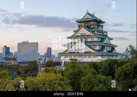 Osaka, Japan - 7. November 2023: Der Hauptturm der Burg Osaka mit seinem mehrstöckigen Dach mit grünen Fliesen und goldenen Akzenten ist in der Abenddämmerung zu sehen, mit modernen Stadtgebäuden und Bäumen im Hintergrund. Stockfoto