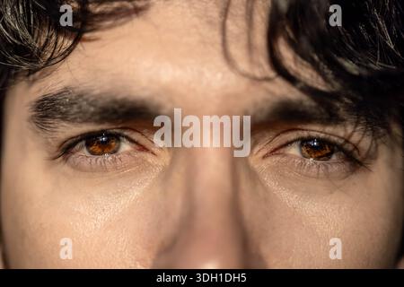Ein extremes Nahbild eines männlichen Gesichts, das sich auf intensiv braune Augen, Wimpern und dicke Augenbrauen konzentriert. Das Bild hebt die Textur der Iris hervor Stockfoto