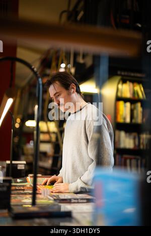 Aufmerksamer Buchliebhaber, der Hörbücher in Pods hört und traditionelle Papierbücher im Geschäft auswählt. Stockfoto
