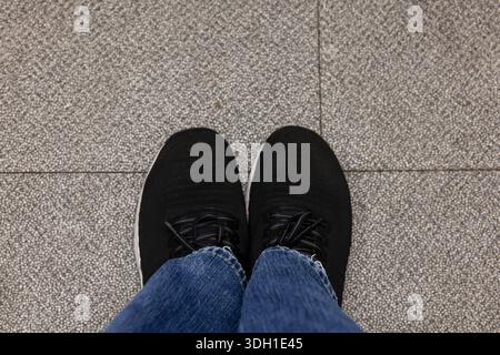 Eine POV-Aufnahme (First-Person-Point of View), die auf einem grauen gesprenkelten ti auf die Füße einer Person blickt, die schwarze Mesh-Laufschuhe und blaue Jeans trägt Stockfoto