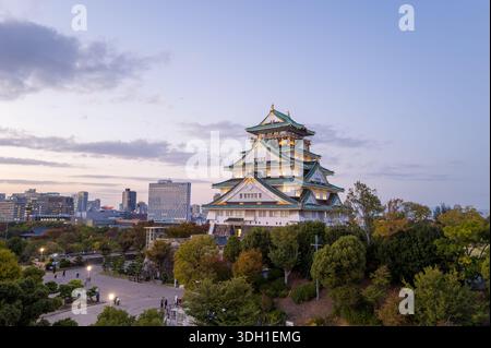 Osaka, Japan - 7. November 2023: Der Hauptturm der Burg Osaka wird am Abend beleuchtet, mit umliegenden Bäumen und modernen Stadtgebäuden im Hintergrund unter teilweise bewölktem Himmel. Stockfoto