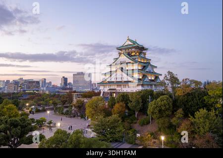 Osaka, Japan - 7. November 2023: Der Hauptturm der Burg Osaka wird in der Abenddämmerung beleuchtet, mit einem umliegenden platz, Bäumen und modernen Stadtgebäuden, die im Hintergrund unter einem teilweise bewölkten Himmel zu sehen sind. Stockfoto