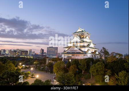 Osaka, Japan - 7. November 2023: Der Hauptturm der Burg Osaka wird in der Abenddämmerung beleuchtet, mit modernen Stadtgebäuden, Bäumen und einem platz, der im Hintergrund unter einem teilweise bewölkten Himmel zu sehen ist. Stockfoto