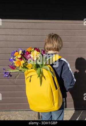 Nicht erkennbarer Junge mit einem hellgelben Rucksack und bunten Blumen darin an sonnigen Tagen. Stockfoto