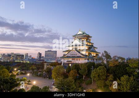 Osaka, Japan - 7. November 2023: Der Hauptturm der Burg Osaka wird in der Abenddämmerung beleuchtet, mit einem umliegenden platz, Bäumen und modernen Stadtgebäuden, die im Hintergrund unter einem teilweise bewölkten Himmel zu sehen sind. Stockfoto