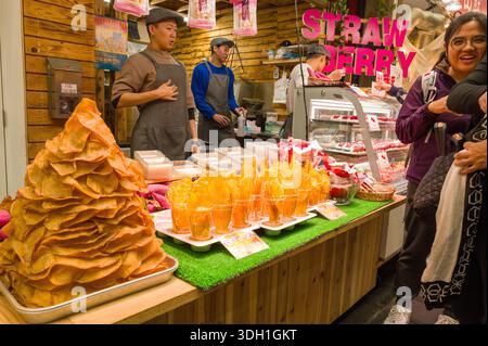 Osaka, Japan - 7. November 2023: Zwei Händler stehen hinter einem Holzstand, der einen großen Haufen Süßkartoffelchips und Tassen mit Chips auf dem Kuromon Ichiba Market zeigt, mit einer gekühlten Kiste Erdbeerdesserts und einem großen rosa „ERDBEERSCHILD“, das im Hintergrund sichtbar ist, wenn die Kunden an der Theke interagieren. Stockfoto