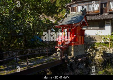 Kinosaki, Japan - 9. November 2023: Ein kleiner roter schintoistischer Schrein steht auf einer Steinbasis neben einem Teich, verbunden durch eine enge Fußgängerbrücke, mit einem traditionellen Holzhaus und Grün im Hintergrund. Stockfoto