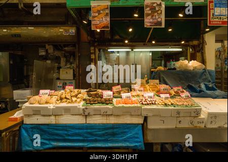 Osaka, Japan - 7. November 2023: An einem Meeresfrüchtestand auf dem Kuromon Ichiba Market werden Schalen mit verschiedenen Muscheln, Austern, Seeigeln und Meeresfrüchtespießen mit handgeschriebenen Preisschildern in japanischen Yen auf Styroporboxen vor einem geschlossenen Ladentisch ausgestellt. Stockfoto