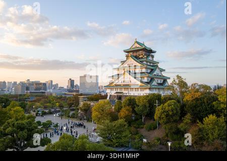 Osaka, Japan - 7. November 2023: Der Hauptturm der Burg Osaka mit seinem mehrstöckigen Dach mit grünen Fliesen und goldenen Akzenten ist in der Abenddämmerung zu sehen, mit Menschen, die sich auf dem platz unten versammeln und der Skyline der Stadt im Hintergrund. Stockfoto