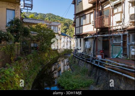 Kinosaki, Japan - 9. November 2023: Ein schmaler Fluss fließt zwischen traditionellen Wohngebäuden mit sichtbaren Rohren und Balkonen, mit Grün und Hügeln im Hintergrund bei klarem Tageslicht. Stockfoto