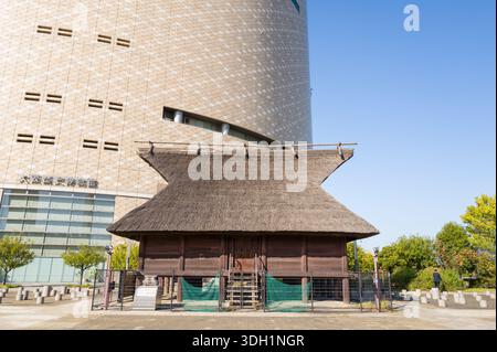Osaka, Japan - 7. November 2023: Ein traditionelles Holzhaus mit Strohdach wird im Freien, umgeben von einem Zaun, vor einem modernen Gebäude mit geometrischen Mustern und japanischen Schildern ausgestellt. Stockfoto