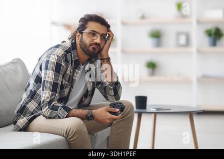 Gelangweilter indischer Kerl, der mit Joystick auf der Couch sitzt Stockfoto