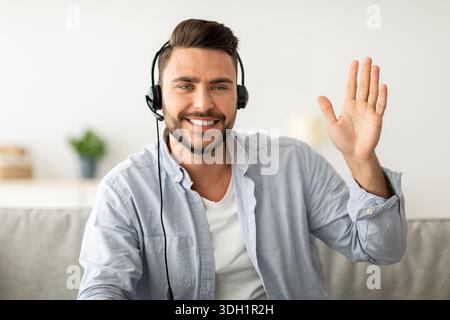 Porträt des Online-Lehrers. Glücklicher, gutaussehender Mann mit Headset, der die Hand zur Webcam winkt und mit Gesten hallo Stockfoto