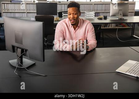 Afroamerikanischer Mann sitzt am schwarzen Schreibtisch mit silbernem Laptop im Büro, in rosa Hemd und spricht Stockfoto