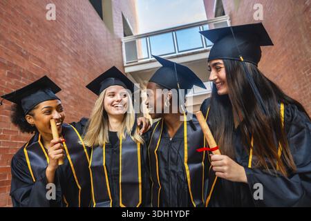 Verschiedene Gruppen von Hochschulabsolventen stehen draußen zusammen, lächeln und lachen, während sie Diplome halten, um ihren Bildungsgang zu feiern Stockfoto
