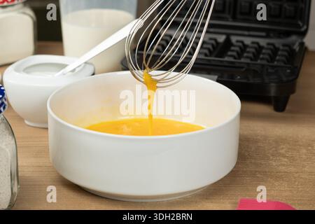 Mit einem Schneebesen die rohen Eier in einer weißen Schüssel zum Backen von Waffeln mischen. Stockfoto