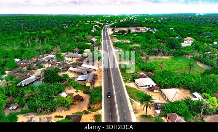 Aus der Vogelperspektive führt ein gerades, graues Straßenband durch eine üppige, grüne Landschaft mit Gebäuden und Bäumen, Ahoada, Rivers State, Nigeria. Stockfoto