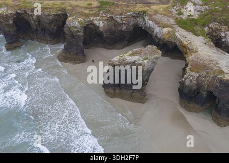 Aus der Vogelperspektive auf die zerklüftete Küste, wo der goldene Sand unter den Schatten der dramatischen Felsformationen auf das türkisfarbene Meer trifft, Ribadeo, Galileien, Stockfoto