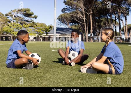 Verschiedene Teenager-Fußballspieler in blauen Hemden sitzen auf dem Fußballfeld und halten 3 Fußballbälle Stockfoto