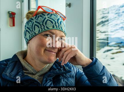 Woman in Winter Gear schaut Kamera an und lächelt mit winterlicher Szene aus dem Fenster Stockfoto