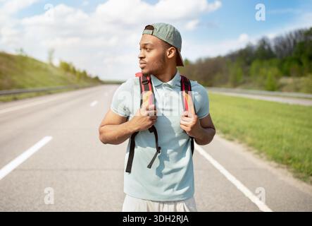 Abenteuer im Sommer. Hübscher junger schwarzer Mann mit Rucksack, der auf einer asphaltierten Straße unterwegs ist und auf der Autobahn unterwegs ist Stockfoto