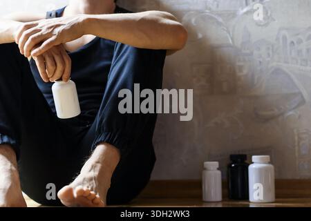 Barfuß-muskulöser Mann in schwarzer Kleidung sitzt auf dem Boden und hängt eine leere weiße Pille-Flasche aus seiner Hand, zusätzliche Flaschen neben ihm auf Holz Stockfoto