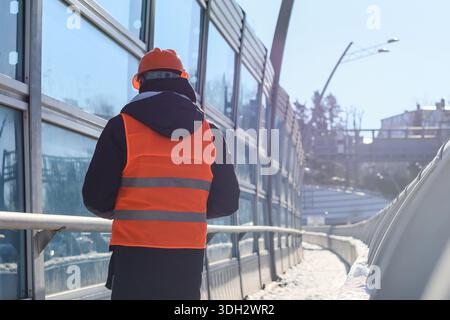 Männlicher Ingenieur in orangefarbener Weste überblickt die Stelle vom erhöhten Weg in verschneite Stadtumgebung Stockfoto