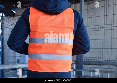 Rückaufnahme des Arbeiters in orangefarbener reflektierender Weste über blauer Jacke, die im Außenbereich mit Industrieumzäunung nach außen zeigt Stockfoto