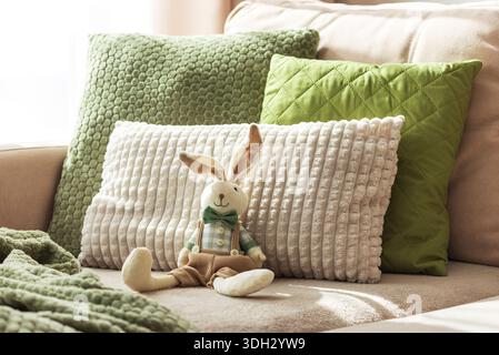 Gemütliches Sofa mit weicher Osterhase-Puppe und strukturierten Kissen im Sonnenlicht Stockfoto