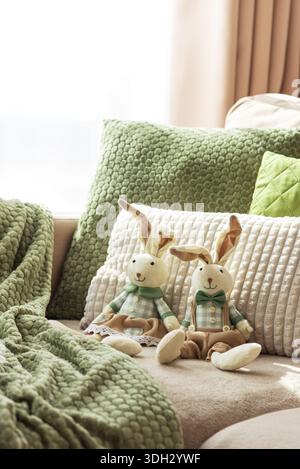 Gemütliches Sofa mit weichen Osterhasen-Puppen und strukturierten Kissen im Sonnenlicht Stockfoto