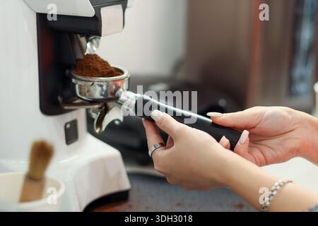 Die Hände des weiblichen Barista halten Portafilter, während frisch gemahlener Kaffee aus der Mahlmaschine in einer professionellen Café-Umgebung ausgegeben wird. Stockfoto