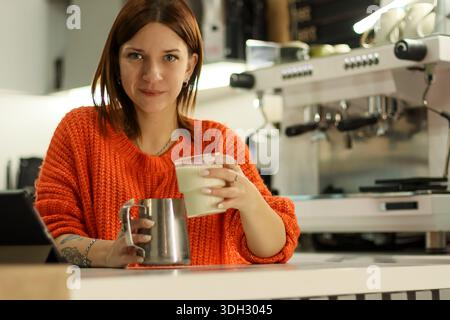 Eine Dame präsentiert eine Tasse Milchgetränk in einer Café-Bar mit sichtbaren Braumaschinen. Stockfoto