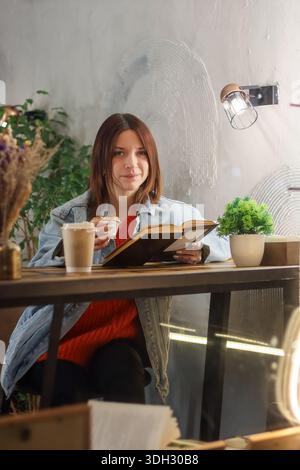 Glückliche junge Frau in Jeansjacke lächelt beim Muffin und beim Lesen von Büchern im gemütlichen Café Stockfoto