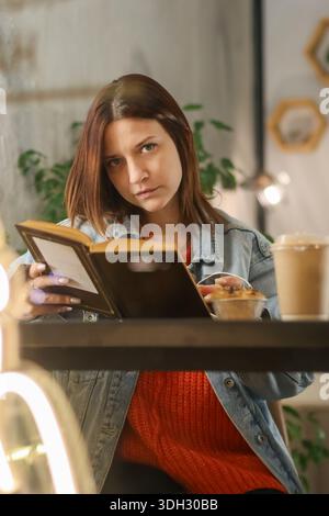 Junge Frau in Jeansjacke liest nachdenklich Buch am Kaffeetisch mit Eiskaffee und Gebäck Stockfoto