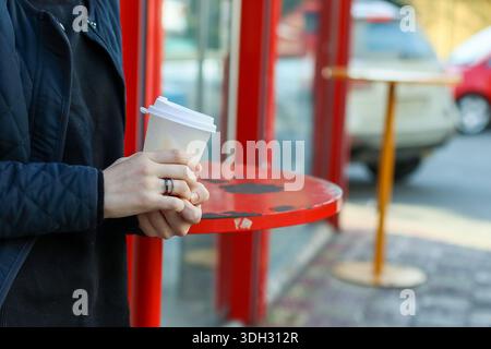Detaillierte Ansicht des weißen Tassen zum Mitnehmen mit weiblicher Hand, mit Deckel auf rotem Tisch in einer Unterkunft für öffentliche Verkehrsmittel in der Stadt Stockfoto