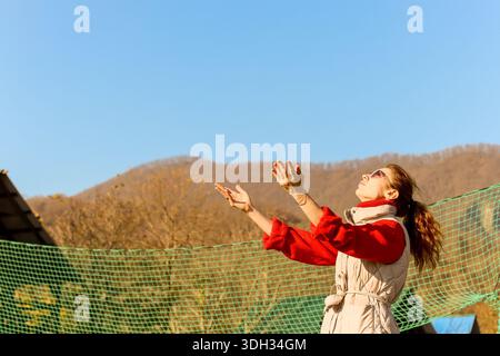 Eine junge Frau in Rot und Beige hebt ihre Hände hoch, von einem Sportnetz in einer sonnigen Landschaft mit Hügeln für Action bereit. Stockfoto
