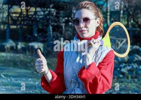 Nahaufnahme einer jungen Frau mit einem Pferdeschwanz, der die Daumen nach oben zeigt, einen farbenfrohen Schläger hält und ein rotes und graues Outfit in einem verschwommenen Outdoor-Hintergrund trägt. Stockfoto