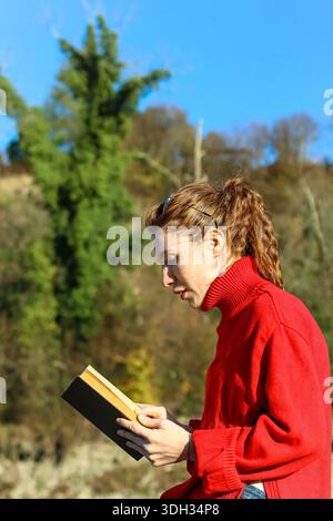Vertikale Aufnahme einer jungen Frau in rotem Pullover, die Buch liest, neben hohem grünen Baum unter hellblauem Himmel steht. Stockfoto