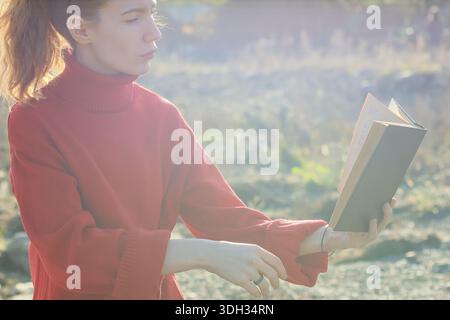 Eine Frau mit Pferdeschwanz in roter Kleidung liest ein offenes Buch in einem natürlichen felsigen Gelände mit trüben Bergen in der Abenddämmerung. Stockfoto