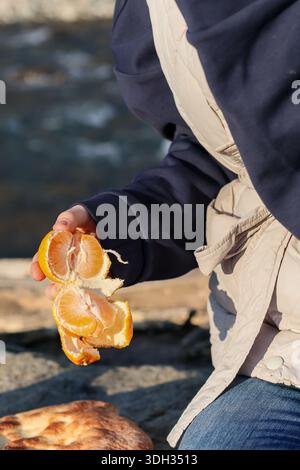 Junge Frau in hellem Mantel, die frisch geschälte Mandarinenorange hält und am ruhigen Herbstfluss steht Stockfoto