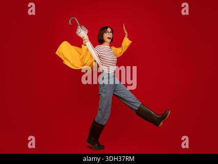 Herbstwetter. Junge Frau in Regenmantel und Gummistiefel kämpfen Wind und gehen über roten Hintergrund Stockfoto