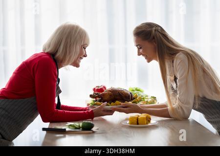 Seitenansicht einer fröhlichen Seniorin mit ihrer jungen, erwachsenen Tochter, die Thanksgiving oder Weihnachts-truthahn in der Küche hält Stockfoto