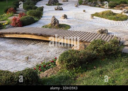 Eine bogenförmige Holzbrücke überspannt elegant einen Zen-Steingarten mit sorgfältig geharkten Sandmustern, strategisch platzierten Steinen, blühenden roten Blumen und l Stockfoto